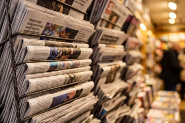 Newspapers stacked in a store showing different sections and headlines in a busy location during the day